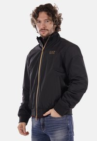 Schwarze Bomberjacke mit hohem Kragen, goldenen Reißverschluss und EA7-Logo. Sie verfügt über elastische Bündchen und einen elastischen Saum, ein glattes Material und zwei Seitentaschen.