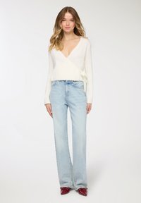 Maglione avvolgente avorio con maniche lunghe e dettaglio annodato, abbinato a jeans a gamba larga azzurri. Scarpe rosse a punta completano il look.