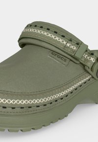 Πράσινο παπούτσι Crocs με διακοσμητικές λευκές και μαύρες ραφές και ρυθμιζόμενο λουράκι στη φτέρνα για ασφαλή εφαρμογή.