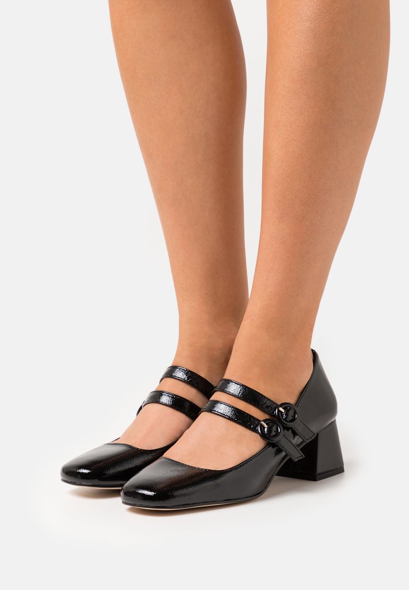 Chaussures Mary Jane en cuir verni noir avec double bride, boucles rondes et talons blocs, portées pieds nus sur un fond clair.