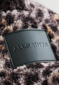 Schwarzes Lederetikett mit geprägtem "ALLSAINTS" auf einem strukturierten, mehrfarbigen, flauschigen Stoff in den Farben Lila, Beige und Braun.