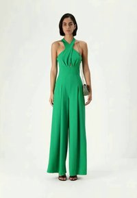 Green wide-leg jumpsuit with a halter neckline and cut-out detail. Made from a smooth fabric, paired with a small patterned handbag and sandals.