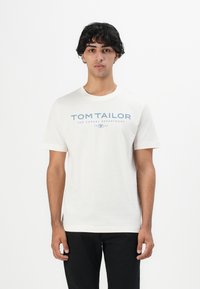 Junger Mann mit lockigem Haar, der ein weißes T-Shirt von "Tom Tailor" und schwarze Hosen trägt, steht vor einem schlichten weißen Hintergrund.