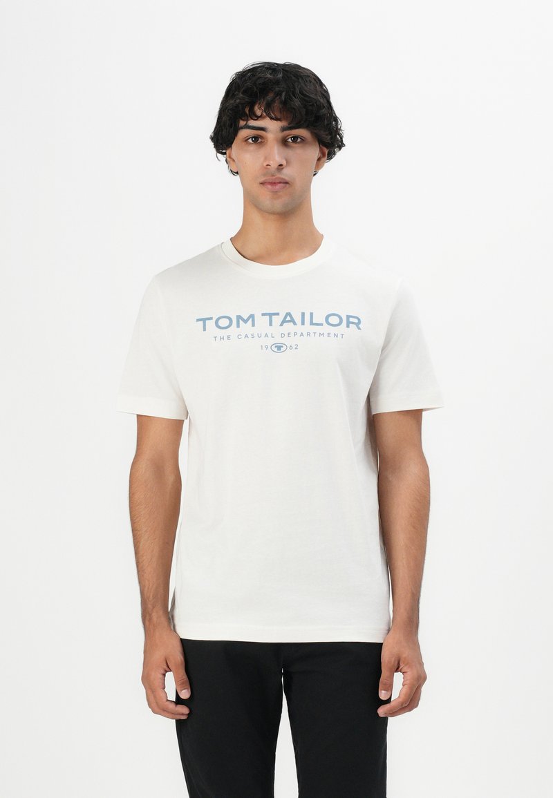Junger Mann mit lockigem Haar, der ein weißes T-Shirt von "Tom Tailor" und schwarze Hosen trägt, steht vor einem schlichten weißen Hintergrund.