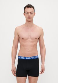Schwarze Boxershorts mit einem blauen Bund, auf dem der Text "BJÖRN BORG" zu sehen ist. Der Stoff wirkt glatt und dehnbar und passt eng am Körper.