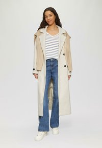 Beige en crème lange jas met grote kraag, gecombineerd met een wit en marineblauw gestreept V-hals shirt, blauwe flare jeans en witte sneakers.