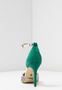 Grön högklackad sko med silverfärgad ankelrem och glittrande detaljer. Mot en enkelfärgad vit bakgrund, som framhäver dess eleganta design.