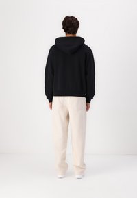 Felpa nera con cappuccio e vestibilità rilassata, con polsini e orlo a coste. Abbinata a pantaloni beige chiaro a vestibilità ampia, dotati di tasche posteriori.