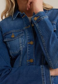 Jeansjacka i medelblått med guldfärgade knappar, fickor och sömmar. Har en kragkrage och långärmar.