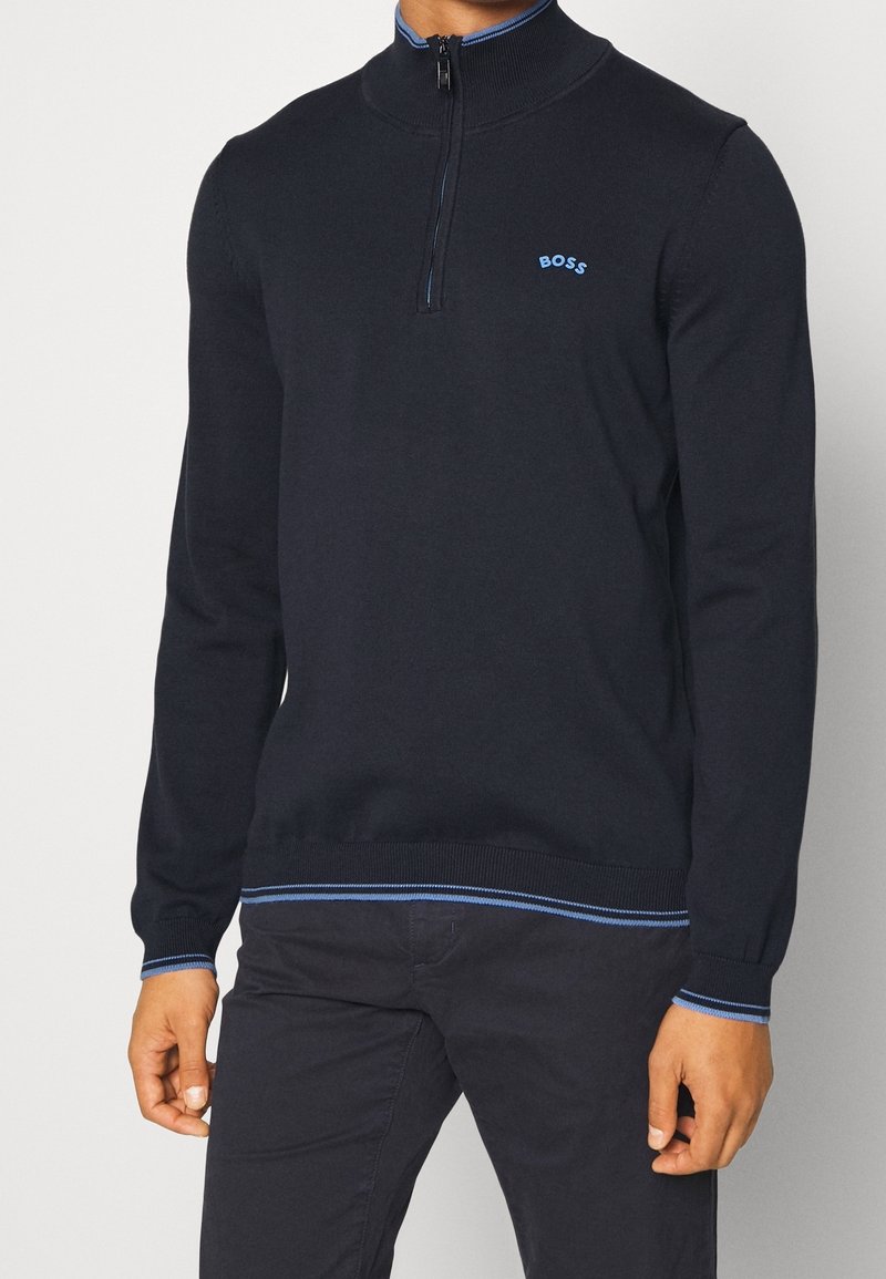 Homme portant un pull BOSS bleu marine à fermeture zippée quart et liseré bleu, avec un pantalon noir, debout devant un fond clair uni.