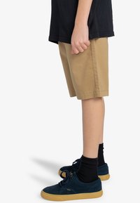 Tan katoenen shorts, knielang, met zijzakken; gedragen met een zwart t-shirt en donkerblauwe suède schoenen met beige zolen.
