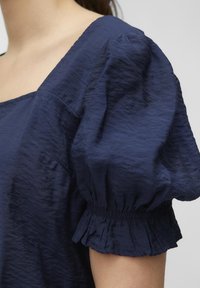 Kvinna iklädd en marinblå blus med puffärmar och elastiska volangmanschetter, visar övre delen av överkroppen och axelområdet.