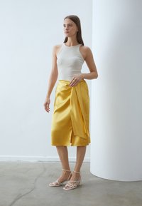 Rok satin kuning dengan desain lilitan dan selubung samping, dipadukan dengan atasan tank beige yang pas. Penampilan ini disempurnakan dengan sandal bertumit blok yang terikat.