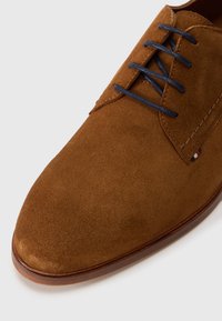 Zapato de ante marrón con punta redonda, cordones en navy y detalle de costura en contraste. Presenta una parte superior suave y texturizada y una suela marrón.