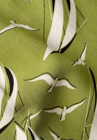 Tessuto di cotone verde con un motivo di uccelli bianchi e accenti neri. La texture è liscia con una leggera lucentezza.