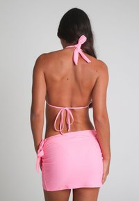 Vrouw met lang, donker haar, gekleed in een roze bikini-top die om de nek en rug is vastgebonden, en een bijpassende roze sarongrok, met haar rug naar de kijker gekeerd.