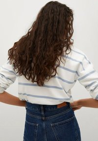 Camicia bianca a maniche lunghe con righe azzurro chiaro, abbinata a jeans di denim vita alta. Cucitura posteriore visibile e toppa in pelle sulla cintura.