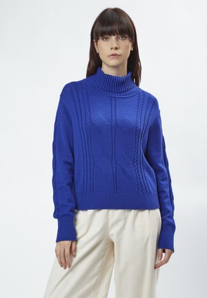 Maglione lavorato a maglia blu cobalto con colletto alto, caratterizzato da motivi a treccia testurizzati e polsini e orlo a coste, abbinato a pantaloni beige chiaro.