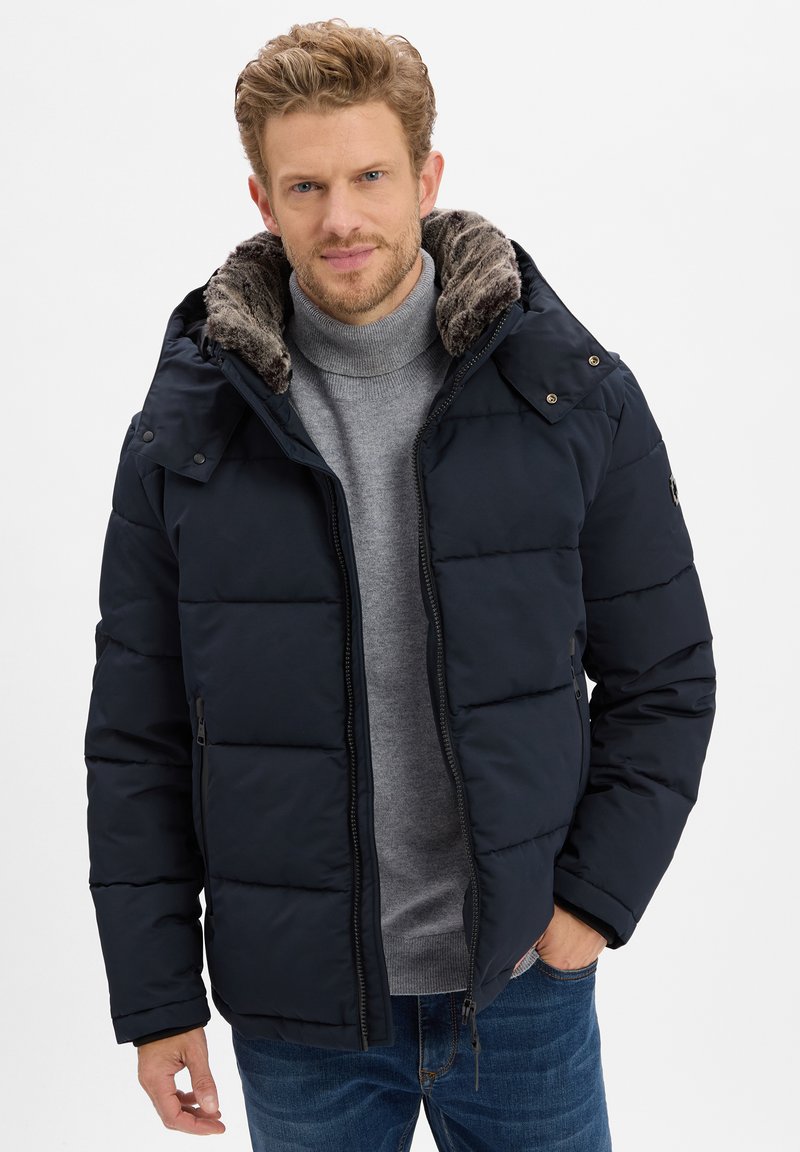 Marineblauwe donzen jas met een bont gevoerde capuchon. Voorzien van een ritssluiting aan de voorkant, zijzakken en een gewatteerde textuur. Gedragen over een grijze coltrui.