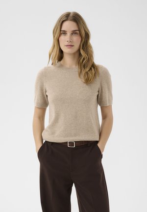 Femme aux cheveux blonds ondulés portant un pull beige à manches courtes et un pantalon marron foncé, les mains dans les poches, debout devant un fond uni.