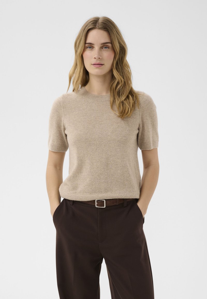 Femme aux cheveux blonds ondulés portant un pull beige à manches courtes et un pantalon marron foncé, les mains dans les poches, debout devant un fond uni.