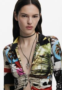 Šarena košulja s kragnom, napola zakopčana, s mješavinom uzoraka na tisku. Crni choker s srebrnim privjeskom u obliku srca.