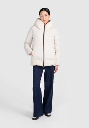 Mujer que lleva una chaqueta blanca acolchada con cuello alto, vaqueros anchos azul oscuro y zapatos negros, de pie frente a un fondo claro y liso.