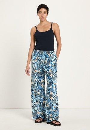 Frau steht, trägt ein schwarzes Tanktop, Hosen mit weitem Bein und blauem Paisley-Muster sowie schwarze Sandalen vor einem einfarbigen hellen Hintergrund.