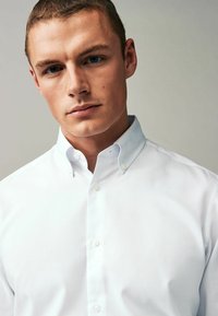 Camisa blanca de botones con cuello extendido, que presenta una textura suave y seis botones en la parte delantera. Diseño limpio, sin patrones ni acentos.