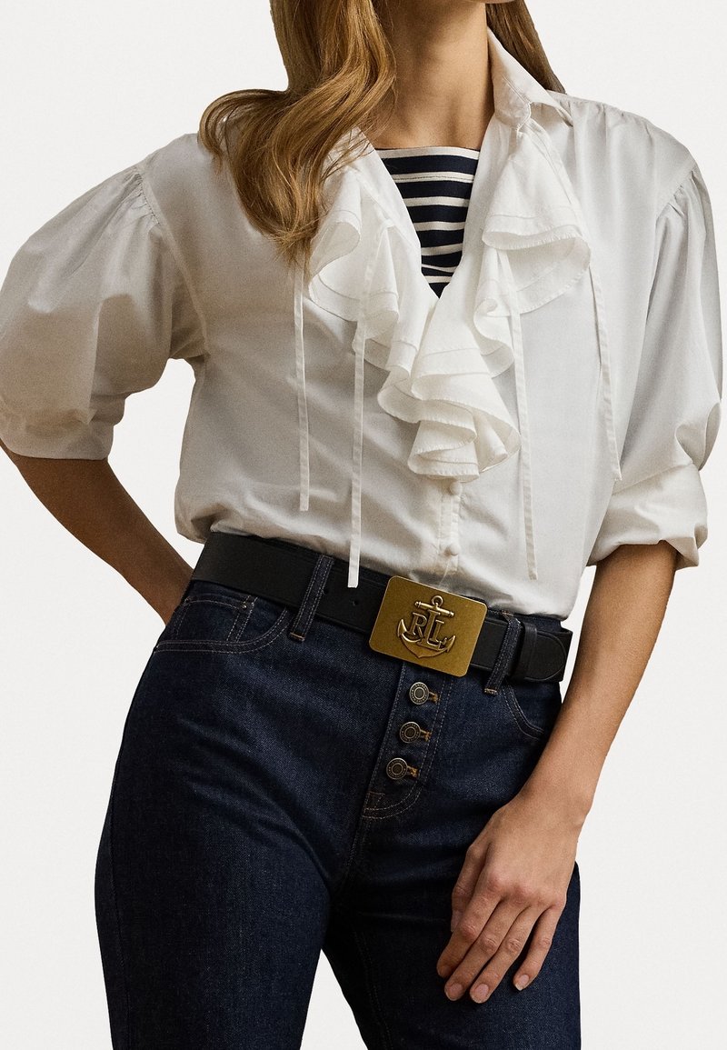 Hvid bluse med flæser og knapper foran, kombineret med mørke denim jeans med høj talje, prydet med en guldfarvet ankerbæltespænde og tre knapdetaljer.