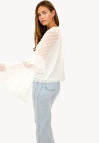 Blouse blanche en tissu texturé, manches à volants et accents floraux, associée à un jean bleu clair. Design simple et élégant.