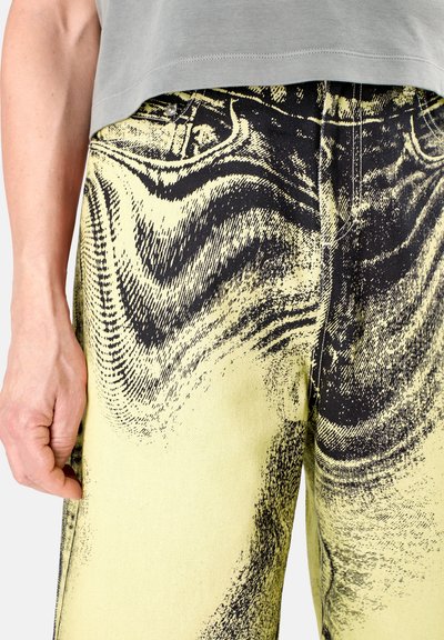 Jeans amarillos con patrones abstractos negros en espiral, con una cintura alta, diseño de cinco bolsillos y detalles de costura visibles.