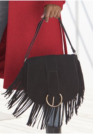 Schwarze Wildleder-Handtasche mit fransenartiger Verzierung, runder Form und einem auffälligen goldenen kreisförmigen Hardware-Akzent. Kombiniert mit einem roten Mantel und Jeans.