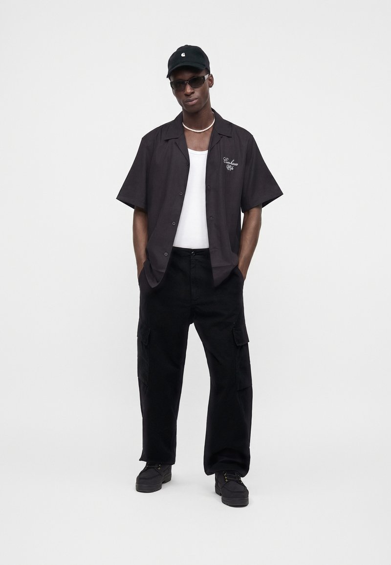 Homme portant une casquette noire, des lunettes de soleil, une chemise noire à manches courtes sur un débardeur blanc, un pantalon cargo noir et des chaussures noires, debout les mains dans les poches.