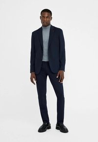 Marineblauwe op maat gemaakte blazer over een grijze coltrui, gecombineerd met smal gesneden marineblauwe broek en zwarte veterschoenen, met een gladde textuur en gestructureerd ontwerp.