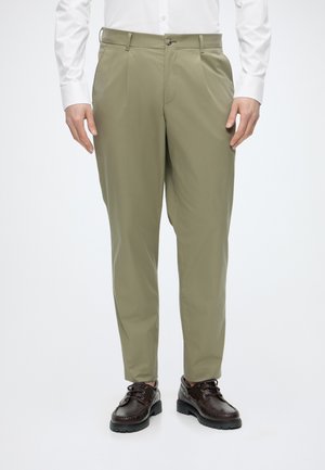 Hombre con pantalones a medida de color verde oliva, camisa blanca de manga larga y zapatos de cuero marrón oscuro con cordones, de pie frente a un fondo blanco.