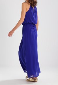 Kungsblå maxiklänning med ett flödande design, ärmlös topp, justerbart dragsko midja och genomskinligt tyg. Gyllene metallsandaler synliga.