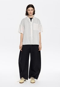 Light-colored, vertically striped button-up shirt with short sleeves and a chest pocket, paired with loose black pants and yellow shoes.