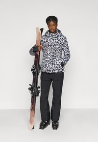 Giacca da sci con stampa leopardata bianca e nera, abbinata a pantaloni da sci neri. Il modello tiene gli sci con gli attacchi, indossa un casco da sci nero e occhiali da sci.