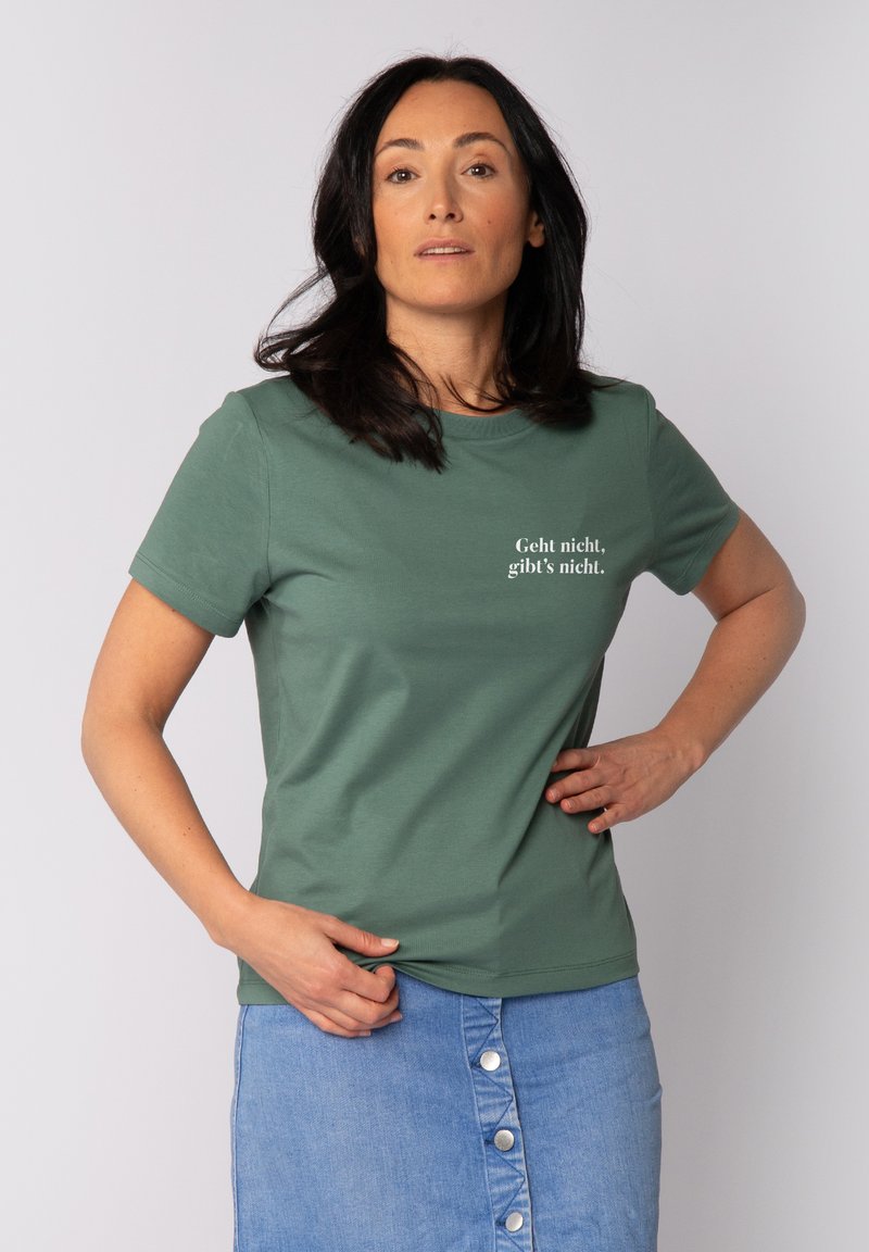 Grünes Baumwoll-T-Shirt mit kurzen Ärmeln, auf der Vorderseite mit dem weißen Text "Geht nicht, gibt’s nicht". Kombiniert mit einem blauen Jeansrock.