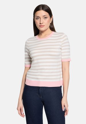 Femme portant un haut à manches courtes beige et blanc rayé avec des bordures roses et un jean bleu foncé, debout devant un fond uni.