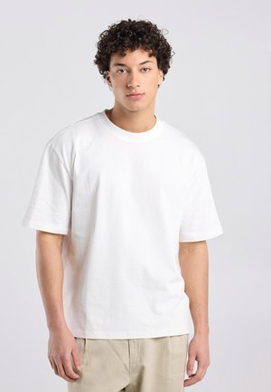 Jonge persoon met krullend haar draagt een effen wit oversized t-shirt en beige broek, staand tegen een lichtgrijze achtergrond.
