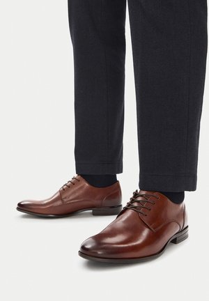 Braune Lederschuhe mit schwarzen Schnürsenkeln, getragen zu dunkler, strukturierter Hose und schwarzen Socken vor hellem Hintergrund.