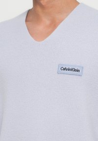 Gai pelēks V veida kakla džemperis, izgatavots no mīksta materiāla ar strukturētu apdari, un ar mazu zilu Calvin Klein logo uz krūšu daļas.