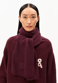 Bordeaux gebreide sjaal met een textuur ribbelpatroon, voorzien van een klein logo patch. Complementair aan een zachte, bordeaux trui.