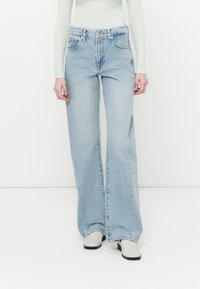 Jean large bleu clair avec un design à cinq poches, texture délavée et couture visible. Porté avec des bottines blanches et un haut à côtes.