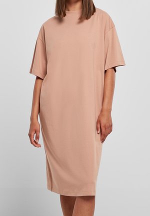 Vrouw draagt een losse, knie-lange beige jurk met korte mouwen, staand tegen een effen lichtgrijze achtergrond.