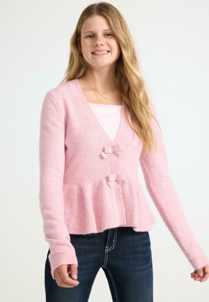 Young woman with long blonde hair wearing a light pink cardigan with bows and dark blue jeans standing against a white background.