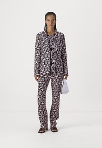 Schwarzer und rosa gemusterter Anzug mit einer langen Jacke und enganliegenden Hosen, die mit floralen Designs versehen sind. Das Model hält eine weiße Handtasche und trägt schwarze Sandalen.