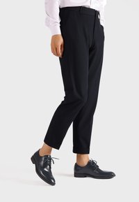 Pantaloni neri con una vestibilità sartoriale, caratterizzati da una texture liscia e risvolti. Abbinati a scarpe stringate in pelle blu scuro lucida.